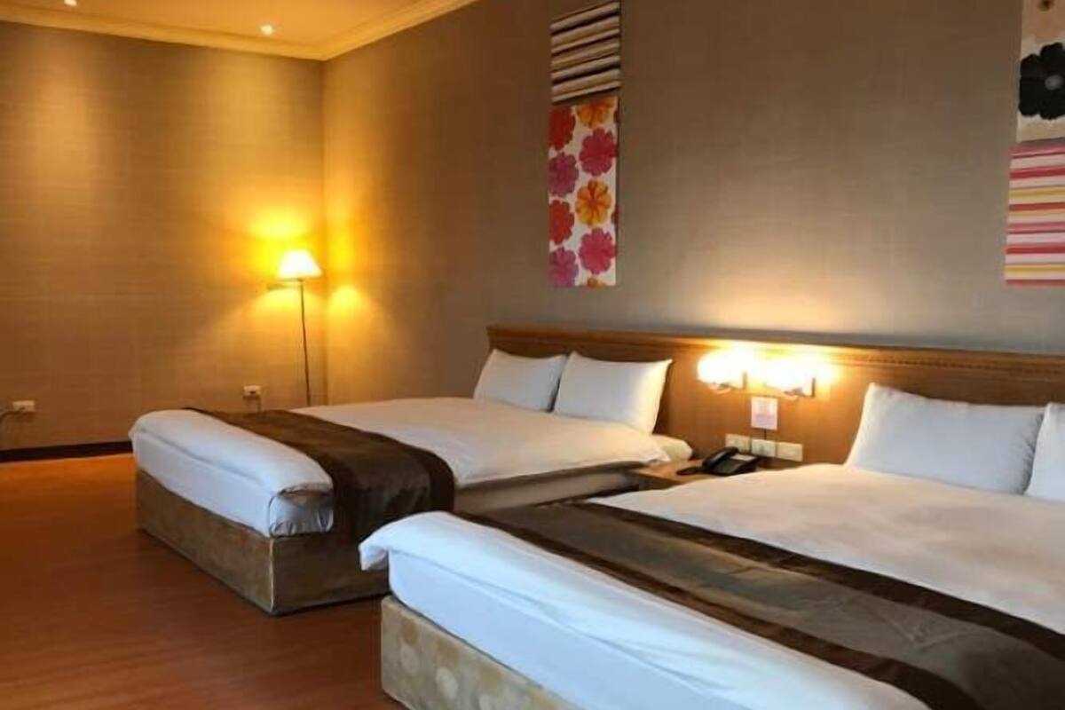 A hotel room with two beds, a wooden floor, a wall-mounted lamp, and a floral artwork.