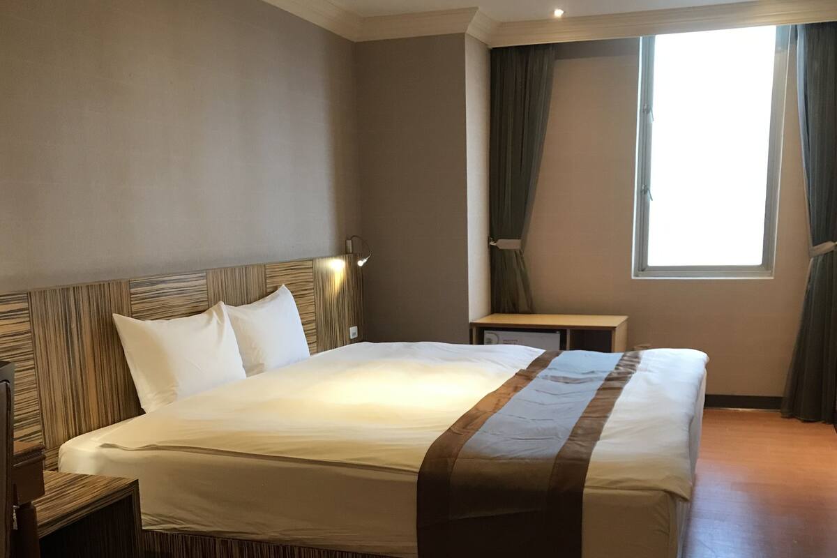 A hotel room with a large bed, a bedside table, a nightstand, a window with curtains, and a wall-mounted lamp.