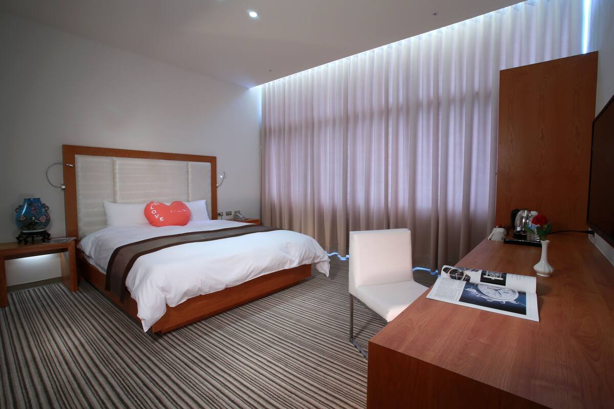 A hotel room with a bed, a desk, a chair, a nightstand, a lamp, a vase with a rose, and a window with curtains.