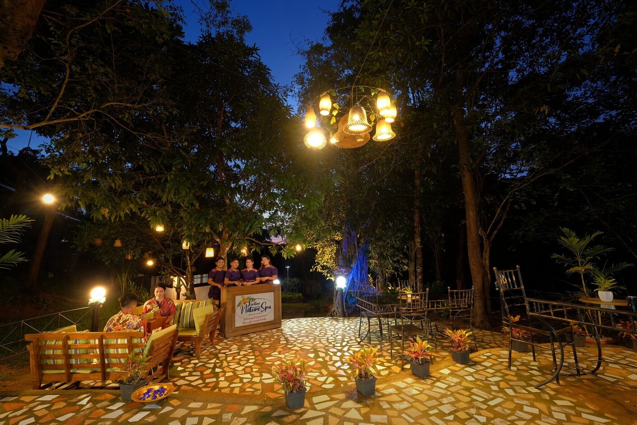 An outdoor seating area with a mosaic floor, wooden furniture, and hanging lights.