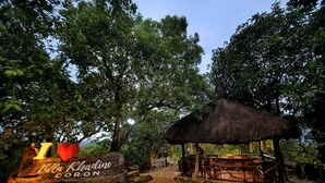 Un espace extérieur rustique, aménagé avec des sièges, et doté d’un toit de chaume, le tout entouré d’une végétation luxuriante. On peut y voir une pancarte en pierre portant l’inscription : « J’adore Villa Khadine CORON ».