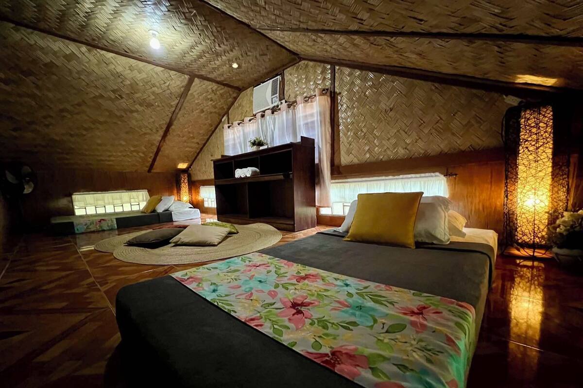 A cozy, rustic room with a high ceiling and wooden beams. It features a bed with a floral bedspread, a seating area with cushions, and a small table.