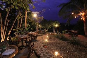Un área de comedor al aire libre con mesas y sillas, rodeada de plantas y palmeras, bajo luces colgantes.