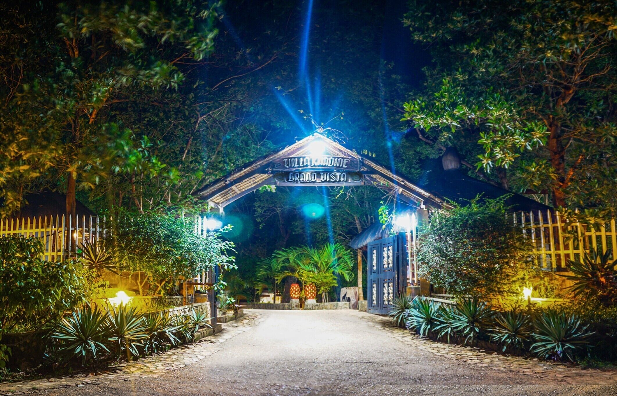 Entrada ben il·luminada a Villa Kandine, amb un rètol, envoltada de vegetació exuberant i un camí pavimentat.