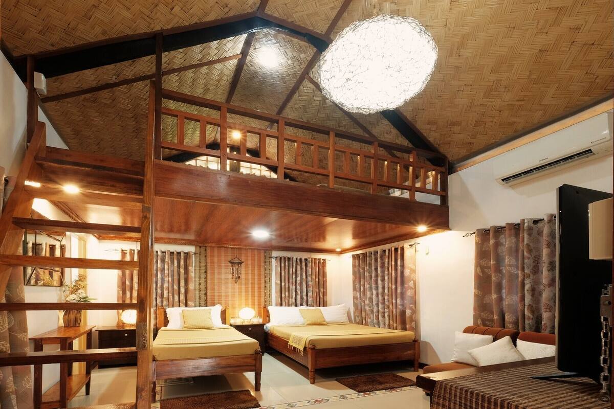 A spacious room with a high ceiling and a wooden staircase leading to an upper level. There are two beds with wooden frames and a small seating area with a table and chairs.