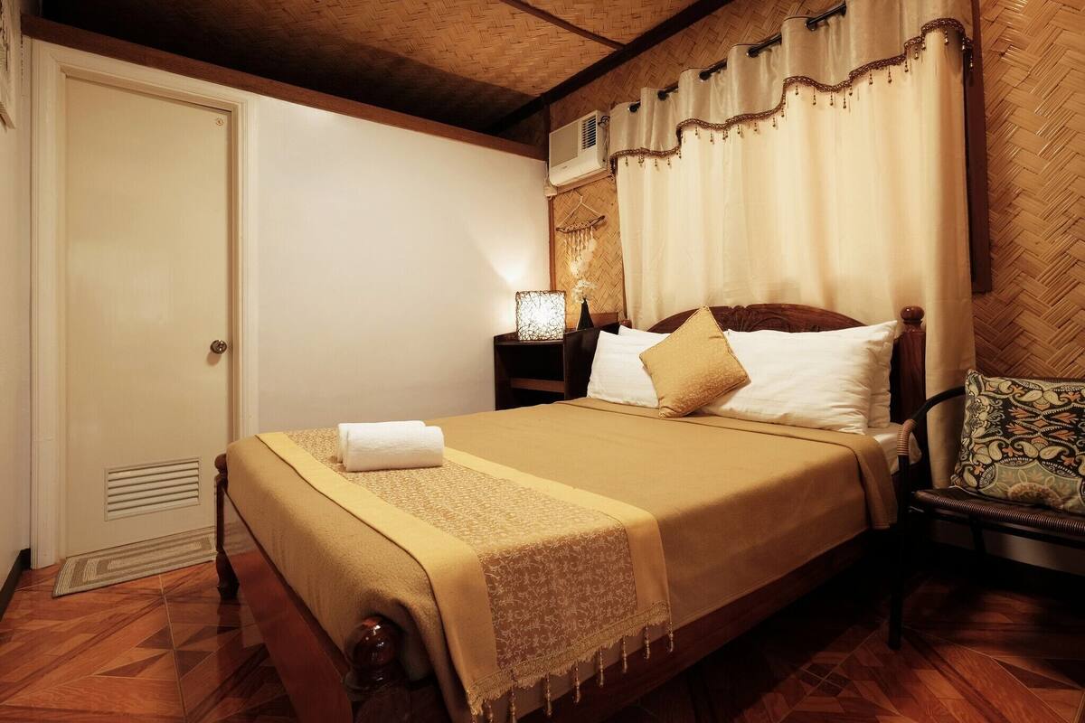 A neatly made bed with a beige bedspread and pillows, a bedside lamp, and a chair with a patterned cushion.