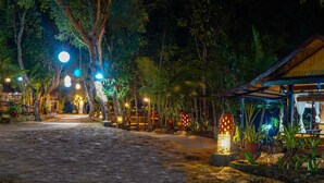 Un espace extérieur aménagé pour la nuit, avec un chemin pavé menant à un bâtiment, entouré d’arbres et éclairé par des lampes suspendues.