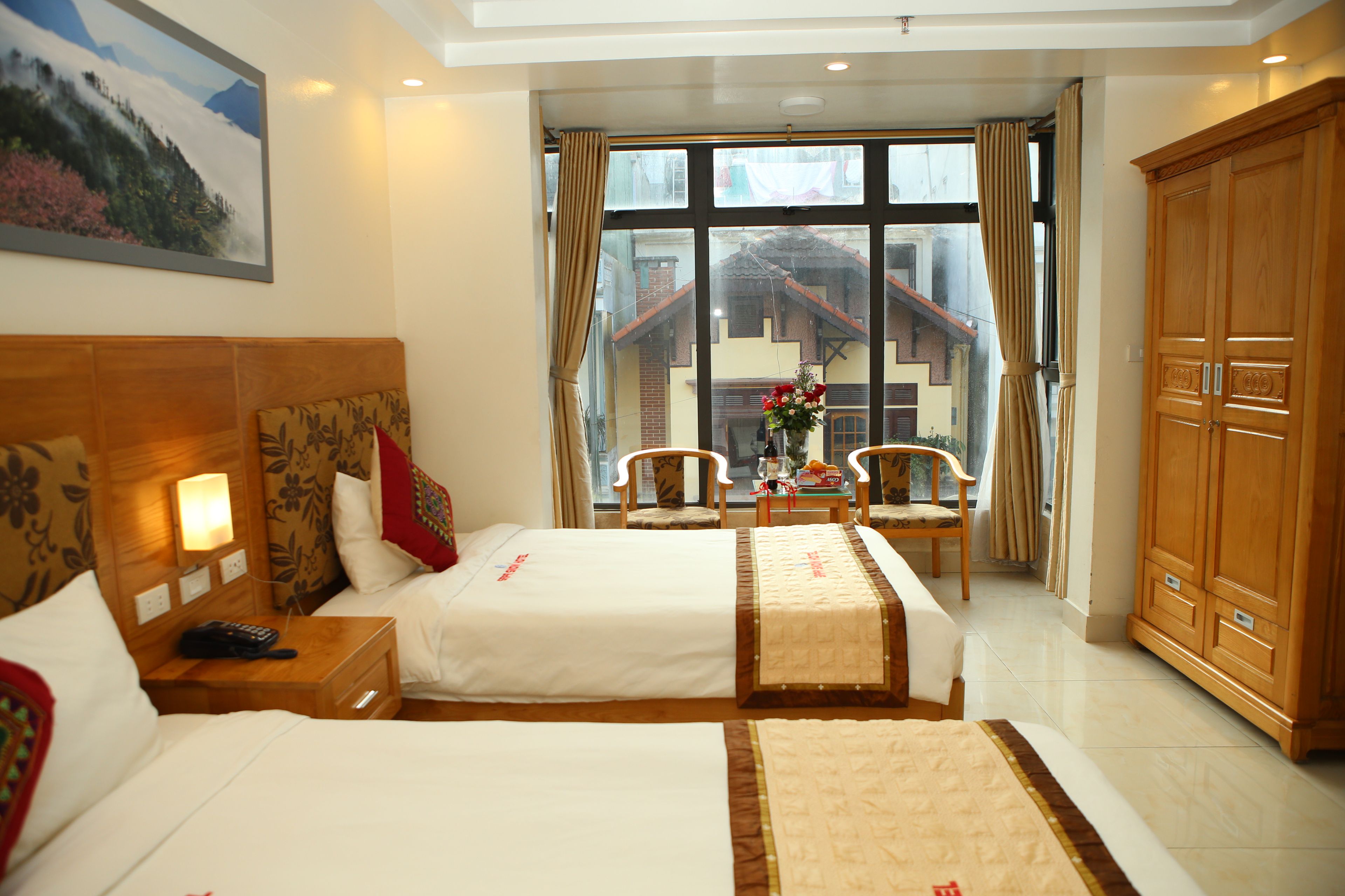 A hotel room with two beds, a wooden wardrobe, a window with a view of a building, and a table with chairs.