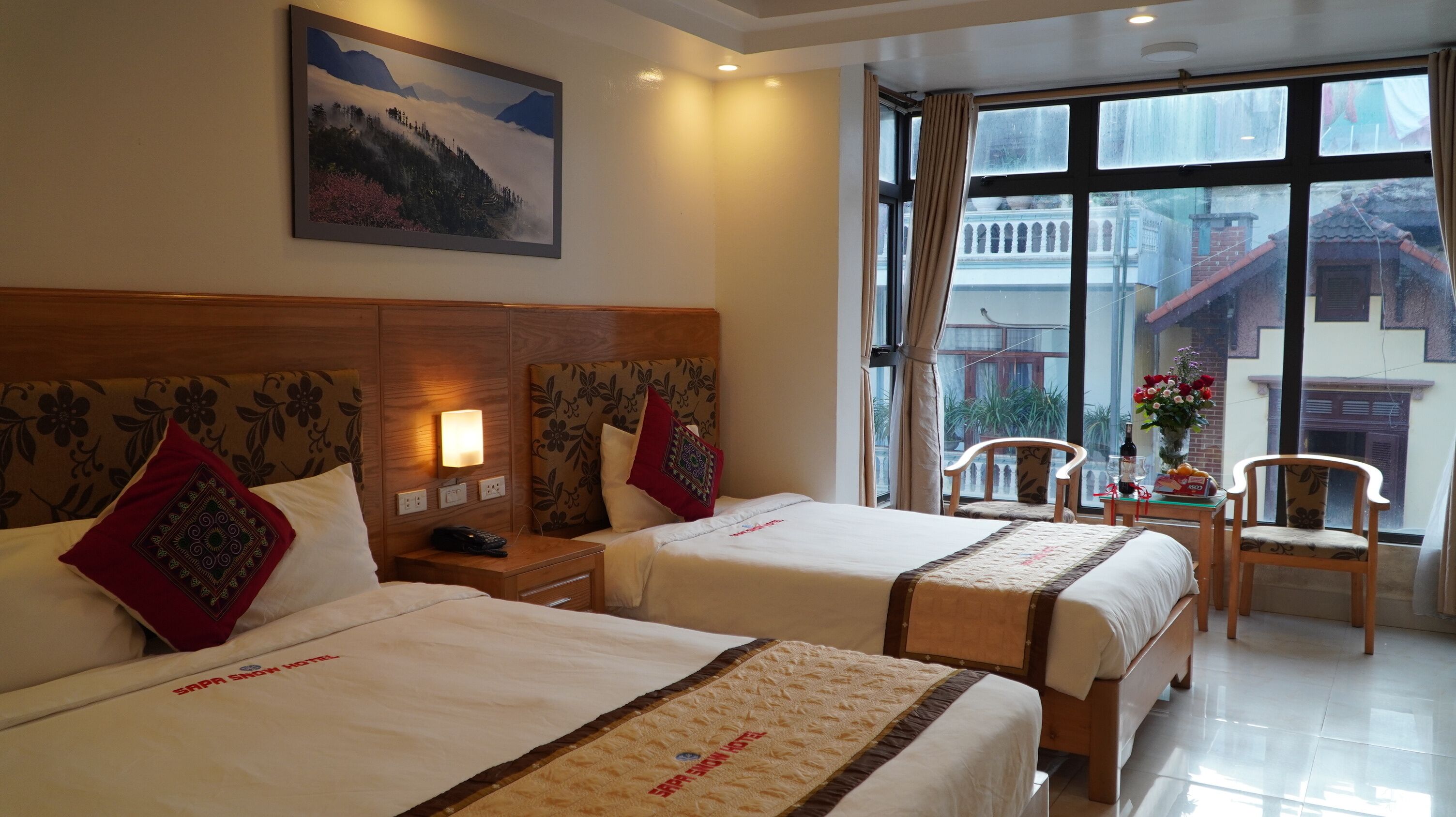 Habitación de hotel con dos camas, una mesita de noche con teléfono, una mesa con un jarrón de flores y una ventana con vistas a edificios.