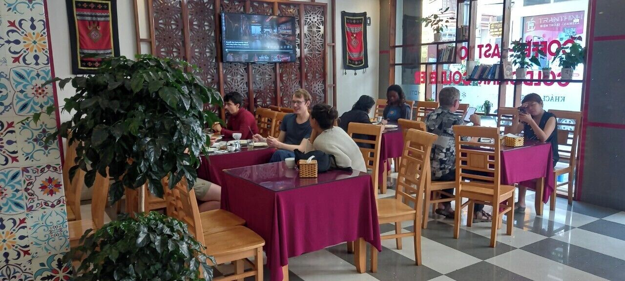 A restaurant with wooden tables and chairs, a large potted plant, and a television screen on the wall.