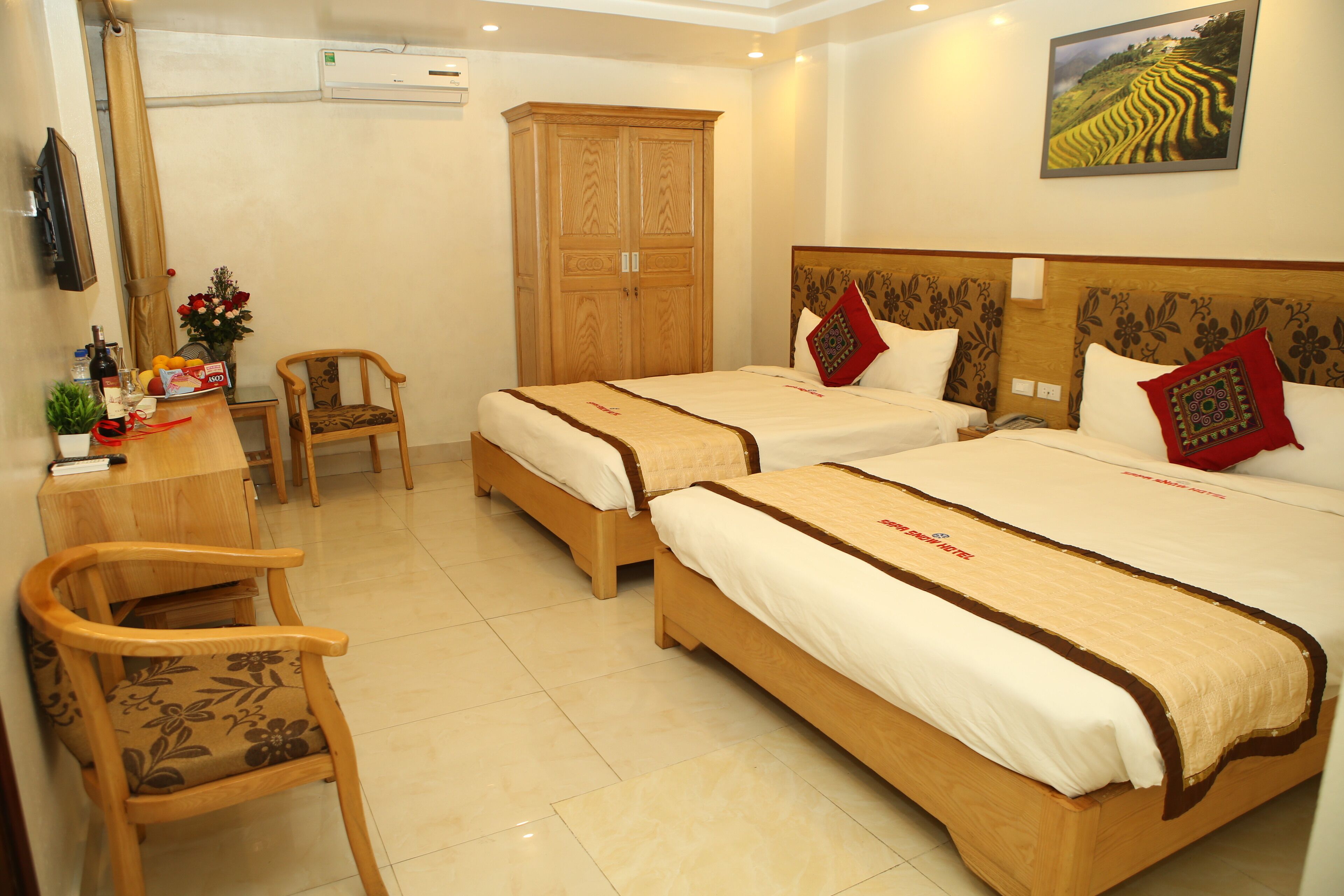 A hotel room with two beds, a wooden wardrobe, a desk with a chair, and a television.