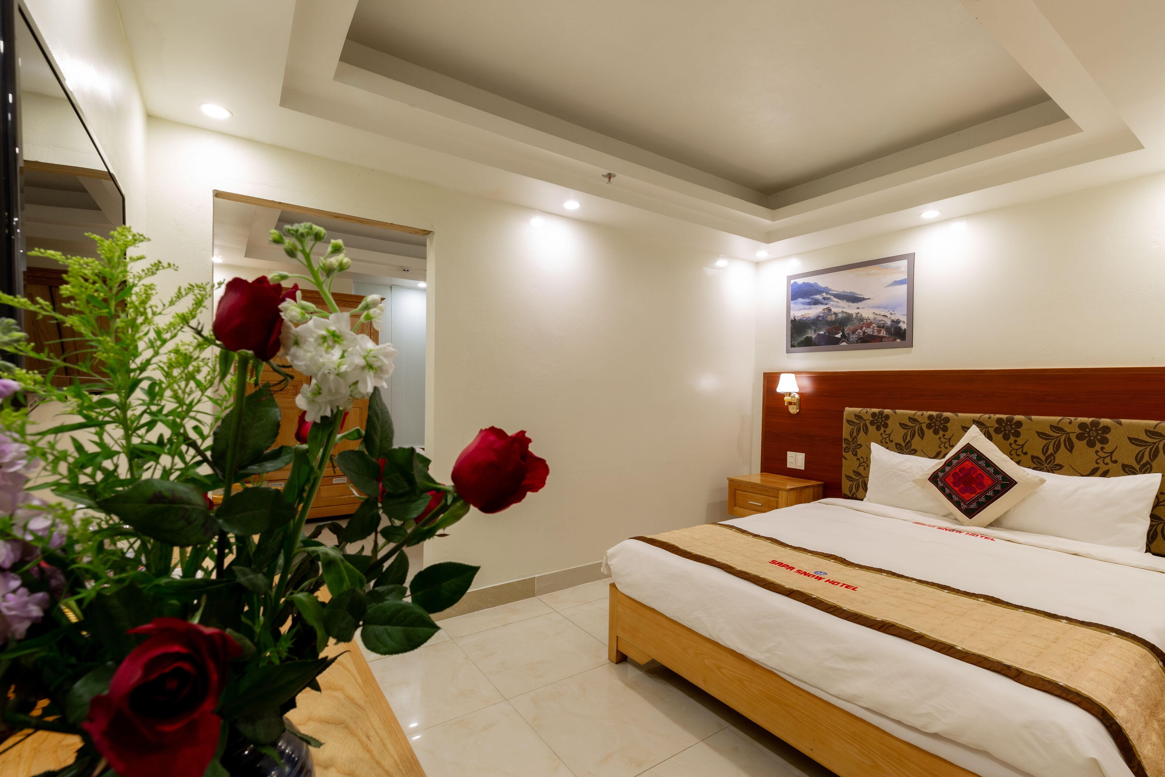 Habitación de hotel con cama de madera, ropa de cama blanca, mesita de noche con lámpara, espejo y decoración floral en la pared.