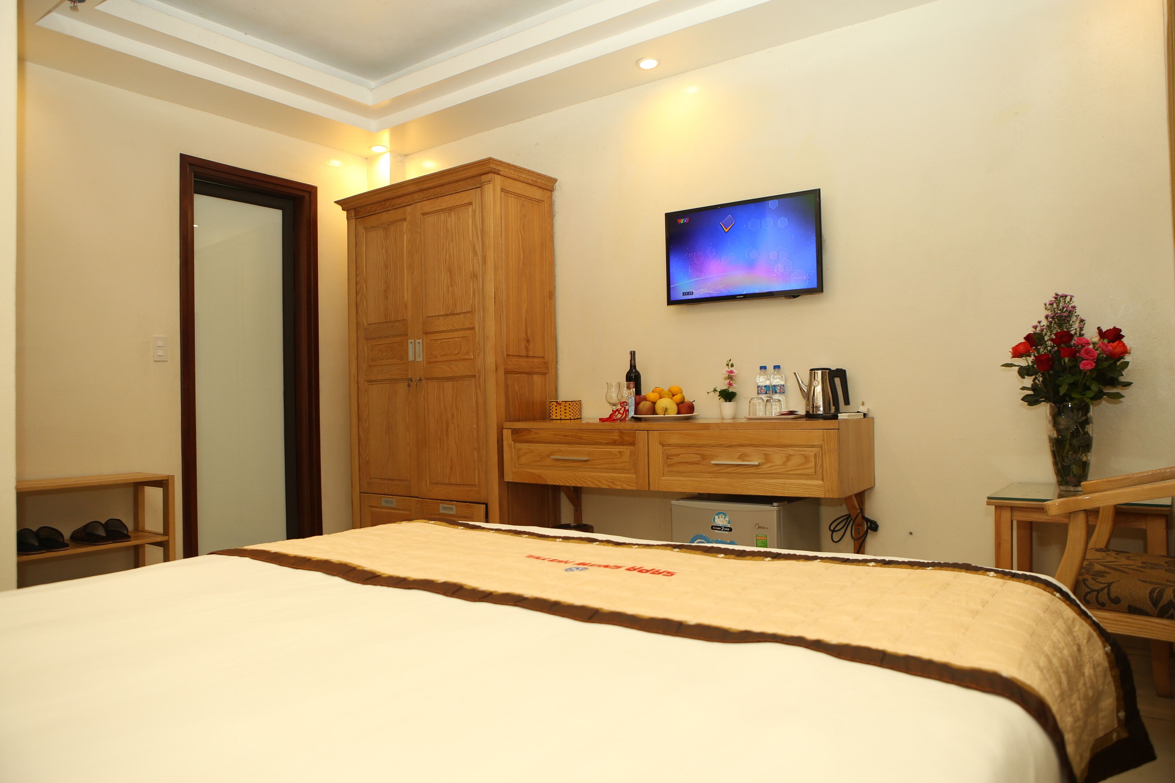 A hotel room with a bed, wooden wardrobe, desk with a flat-screen TV, and a small table with a vase of flowers.