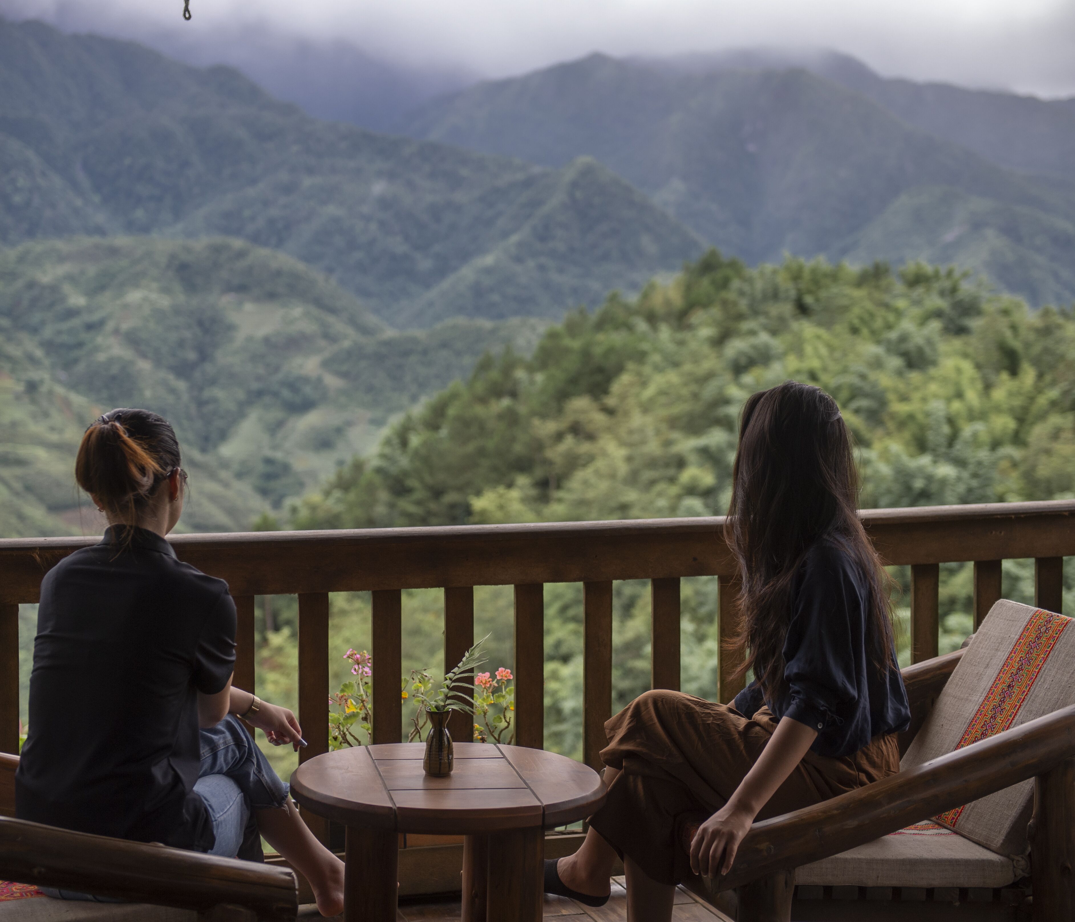 Deux personnes sont assises sur une terrasse en bois, devant une table et un vase de fleurs, et elles ont une vue sur un paysage montagneux.