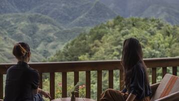 Dos personas sentadas en una terraza de madera con una mesa y un jarrón de flores, con vistas a un paisaje montañoso.