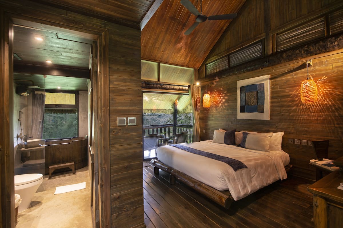 Une chambre à coucher dans une cabane en bois, avec un lit, un ventilateur de plafond, une fenêtre et une vue sur l’extérieur.