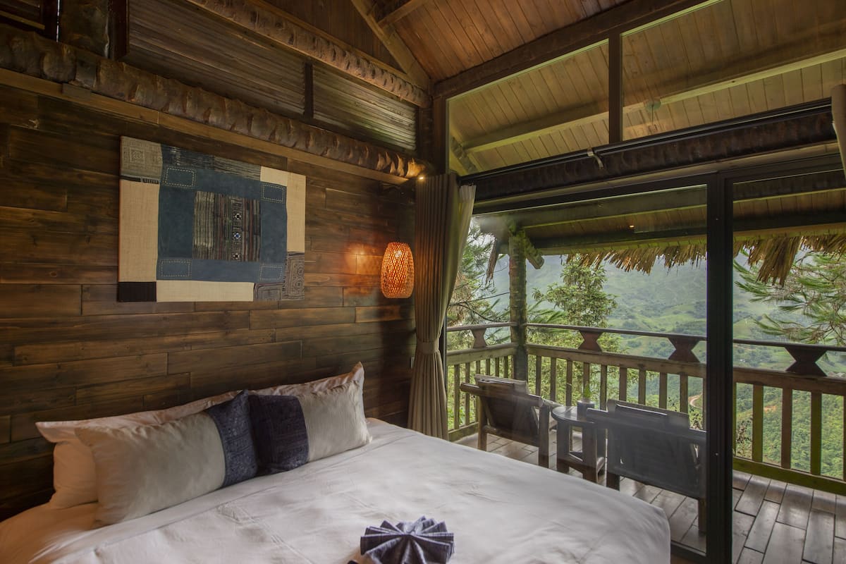 Une pièce de cabane en bois comprenant un lit, un luminaire suspendu et une vue sur un paysage luxuriant, visible à travers une grande fenêtre.