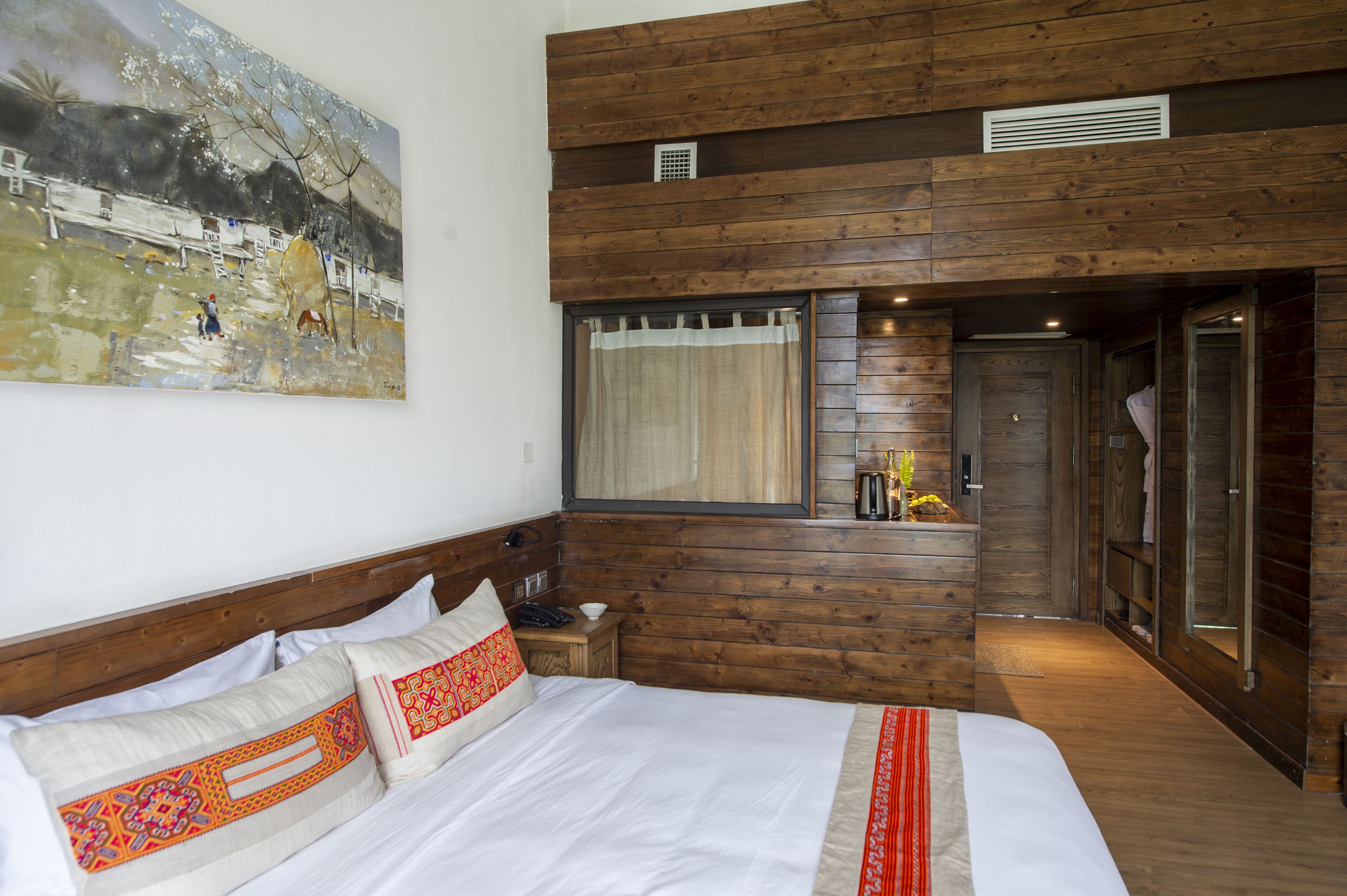 A bedroom with a large bed, wooden walls, a window with curtains, a small table with a phone, and a painting on the wall.