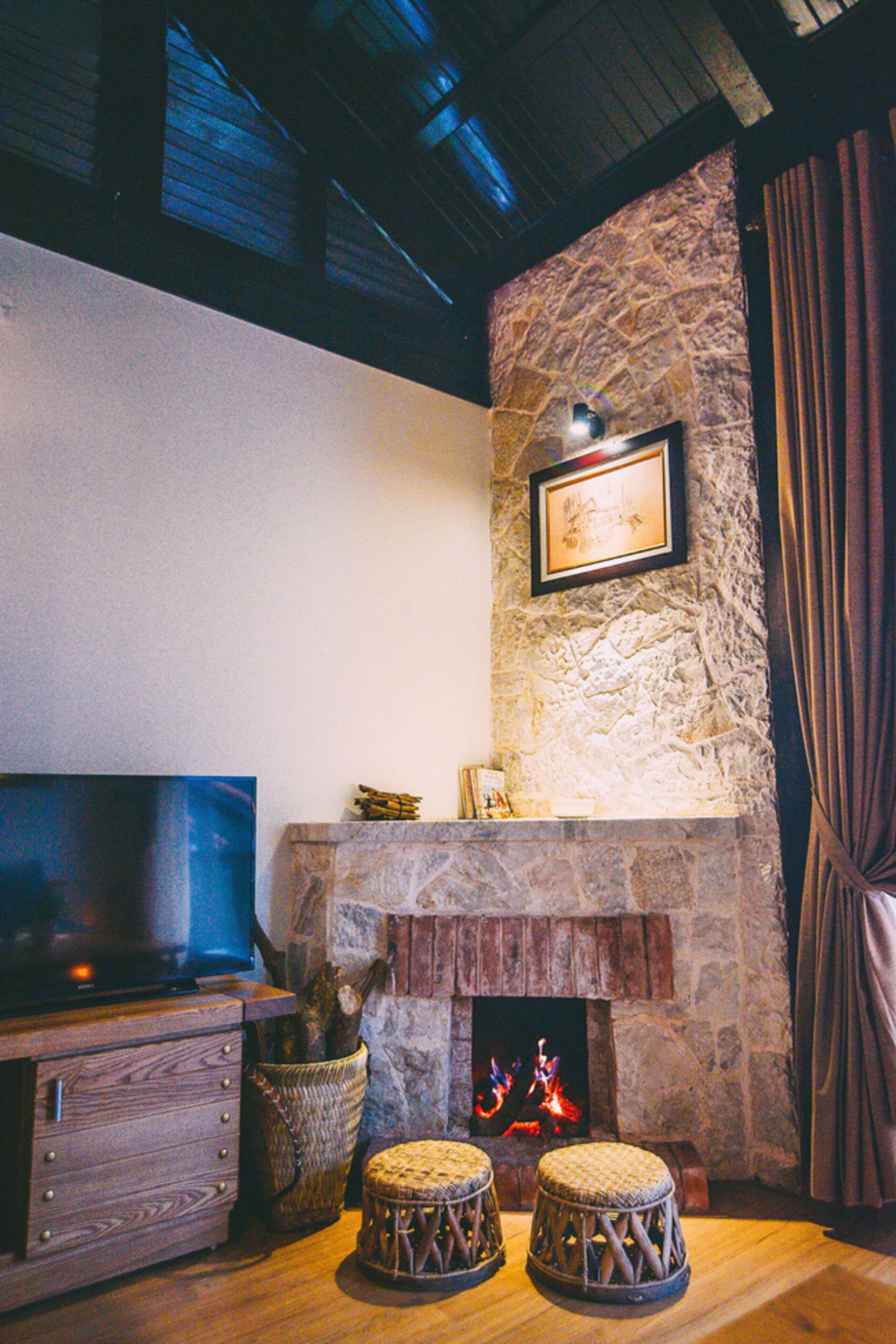 A stone fireplace with a framed picture above it, a television on a wooden cabinet to the left, and two woven stools in front.