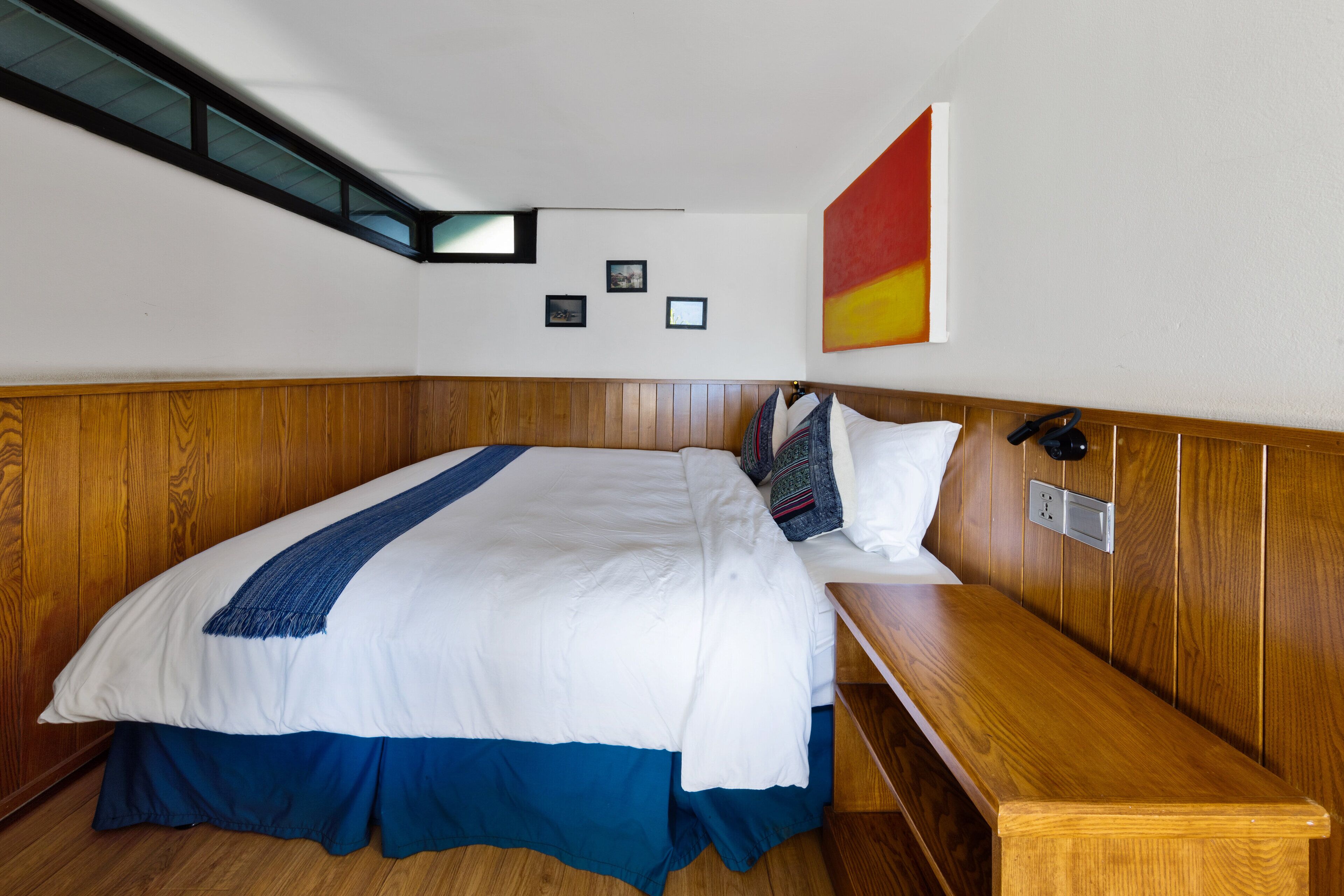 A hotel room with a wooden headboard, a large bed with a blue and white bedspread, a bench at the foot of the bed, and a wall-mounted phone.