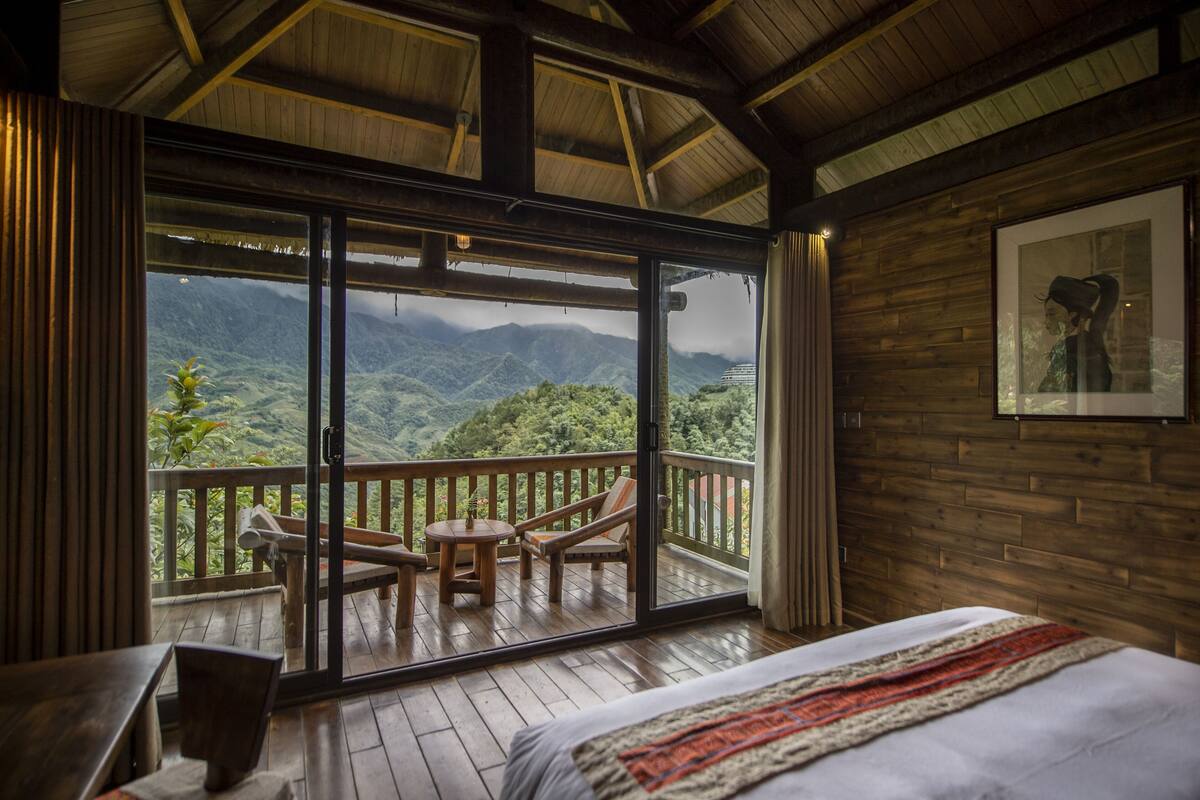 Une chambre dotée d’une grande porte-fenêtre coulissante donnant sur un balcon aménagé avec des meubles en bois et offrant une vue sur les montagnes.