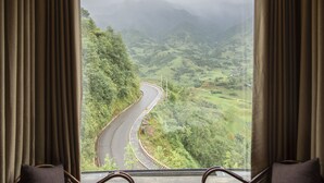 A room with a large window overlooking a winding road and lush green hills. There are two wicker chairs and a small round table with a lamp. - Sapa Jade Hill Resort & Spa (Sa Pa)