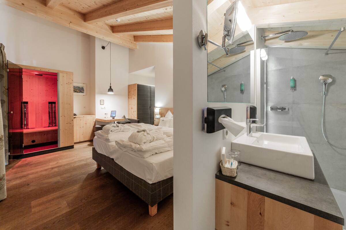 A modern bedroom with a bed, wooden floor, and a red-lit room in the background. It also features a bathroom with a large mirror, sink, and shower.