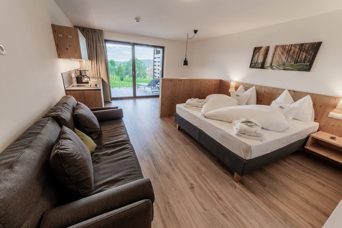 A modern hotel room with a large bed, a sofa, a desk with a coffee maker, and a sliding glass door leading to a balcony with a view of greenery and buildings.