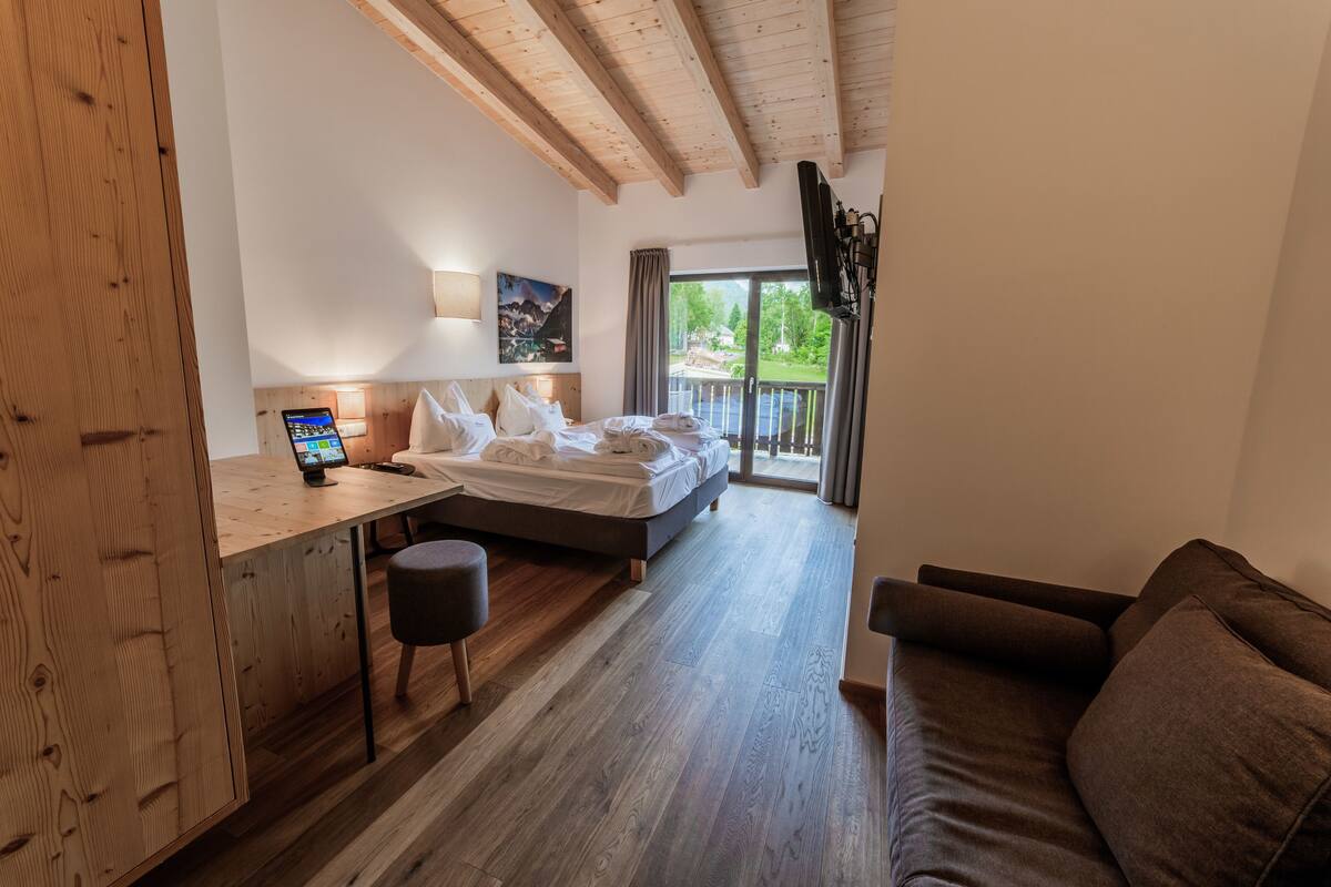 A modern hotel room with a wooden ceiling, a bed, a desk with a computer, a couch, and a large window with a view of greenery.