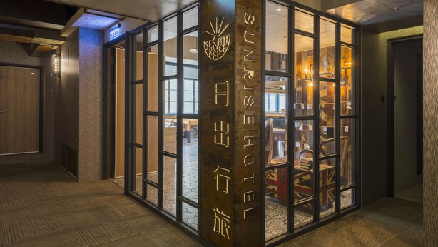 L’entrée d’un hôtel, avec une enseigne indiquant « SUNRISE HOTEL », et on peut apercevoir l’intérieur, qui est décoré de meubles en bois et comprend une cheminée.