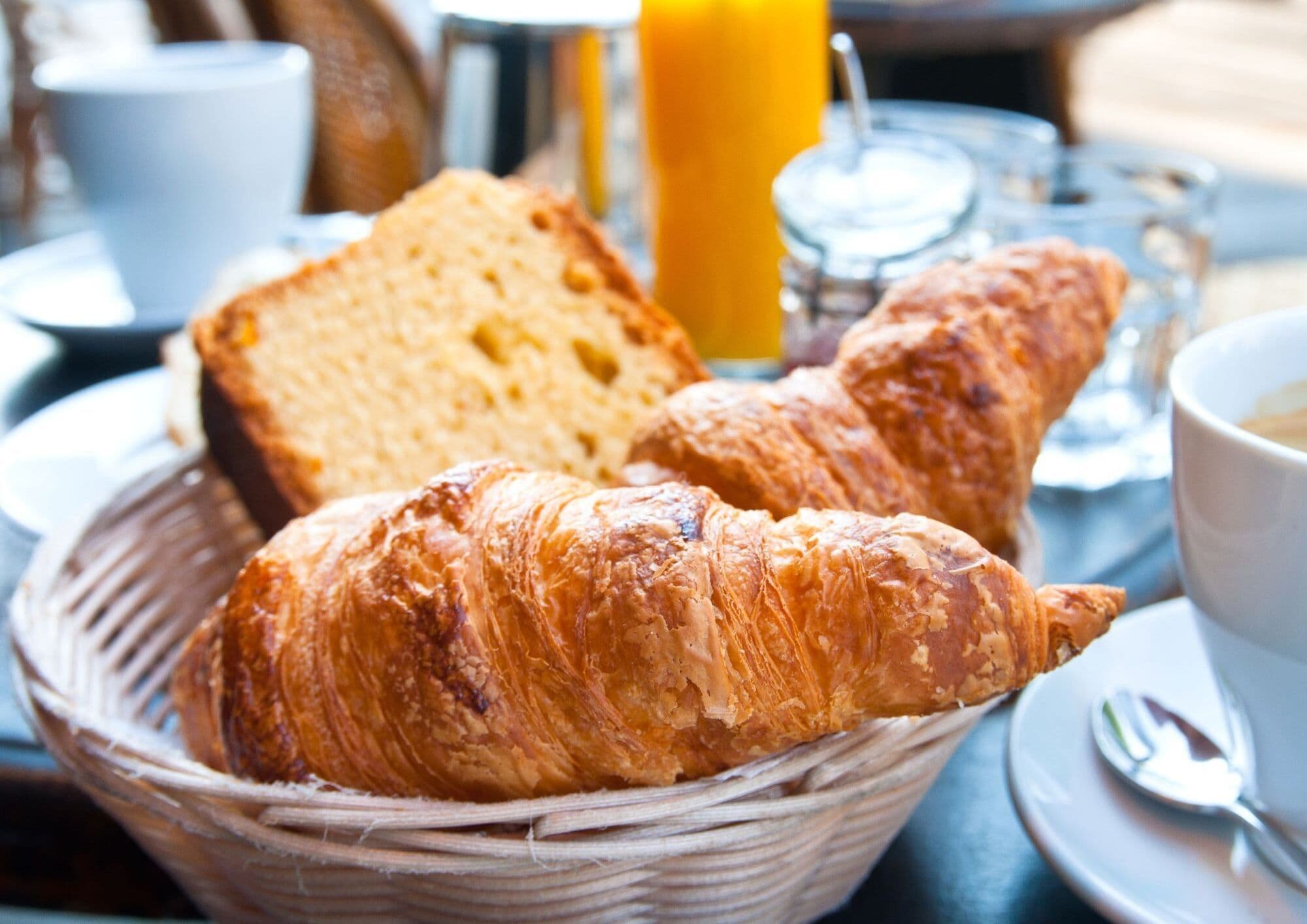 Een mand met croissants en een sneetje brood op een tafel, met een glas jus d'orange en een kop koffie.