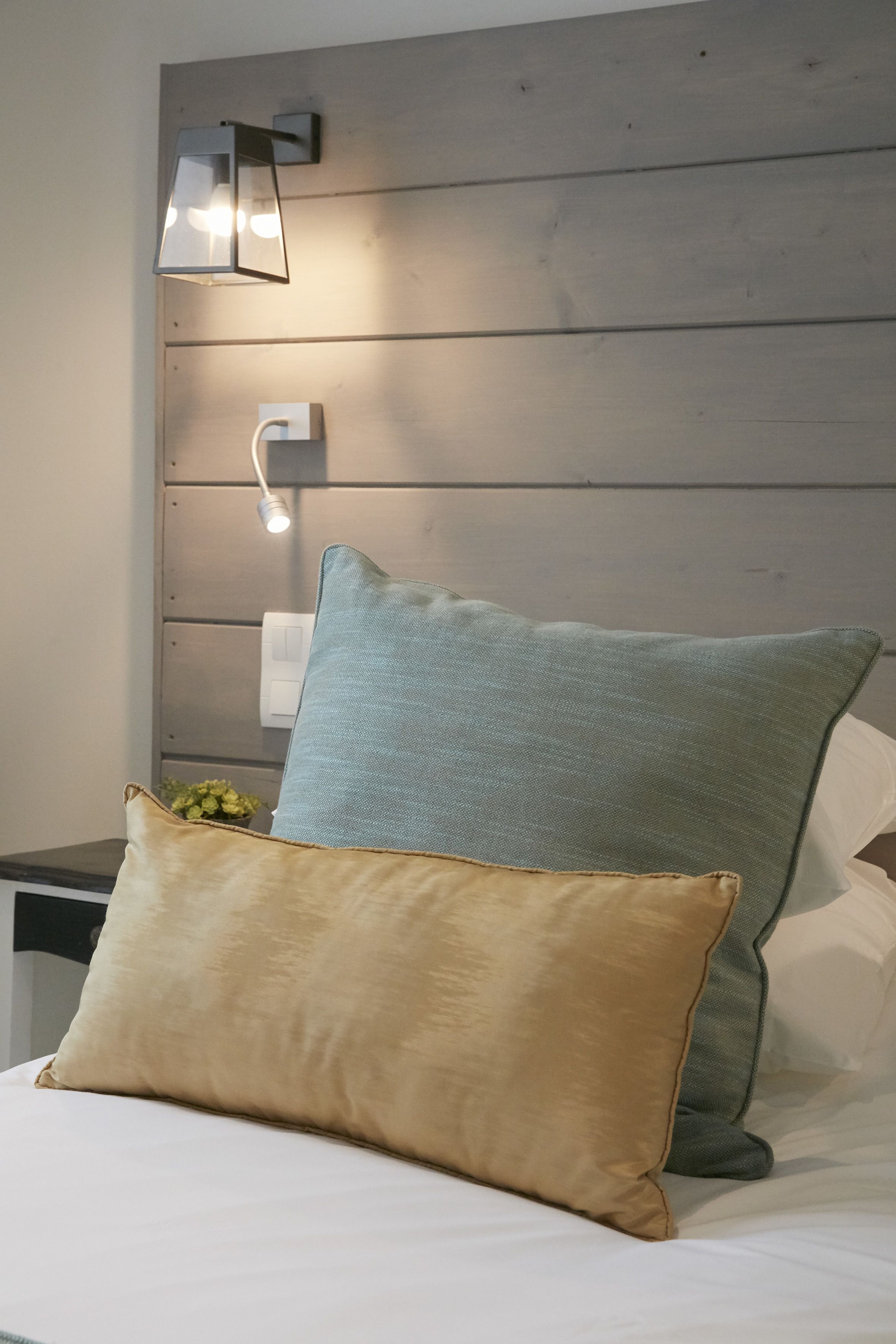 Una cama bien hecha con dos almohadas, una lámpara de pared y una planta pequeña en la mesita de noche.