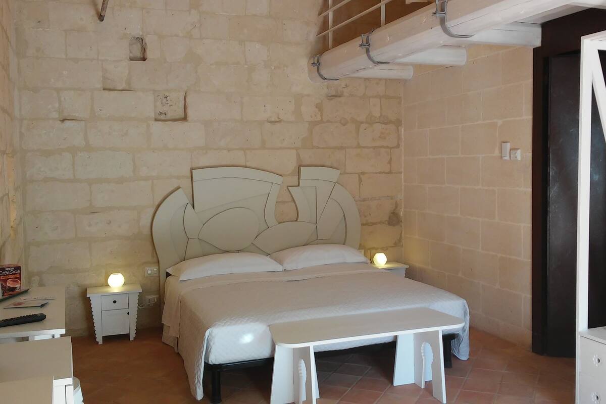 A bedroom with a stone wall, a bed with a white headboard, bedside tables with lamps, and a wardrobe.