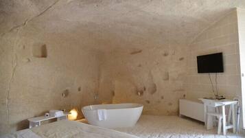 A room with a stone wall, a white bathtub, a bed, a desk, and a flat-screen TV mounted on the wall.