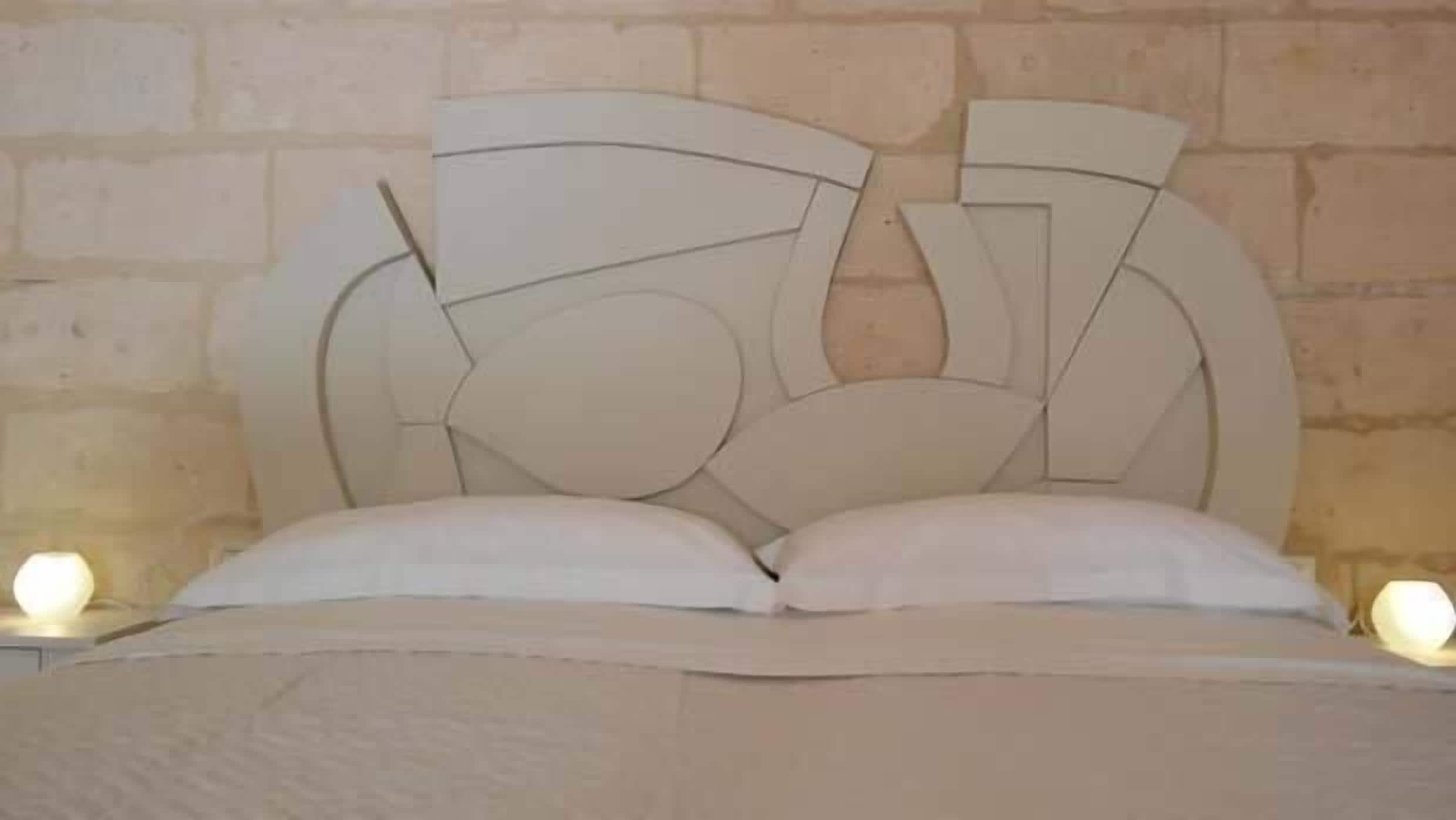 A bed with a unique geometric headboard design, a plain wall, and a nightstand with a lamp.