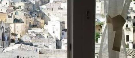 A view from a window with sheer curtains, overlooking a densely packed urban area with buildings of varying heights and architectural styles.