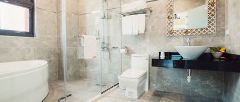 Un baño moderno con ducha envuelta en vidrio, inodoro blanco, una bañera blanca grande y encimera negra con lavamanos blanco y un cuenco decorativo.