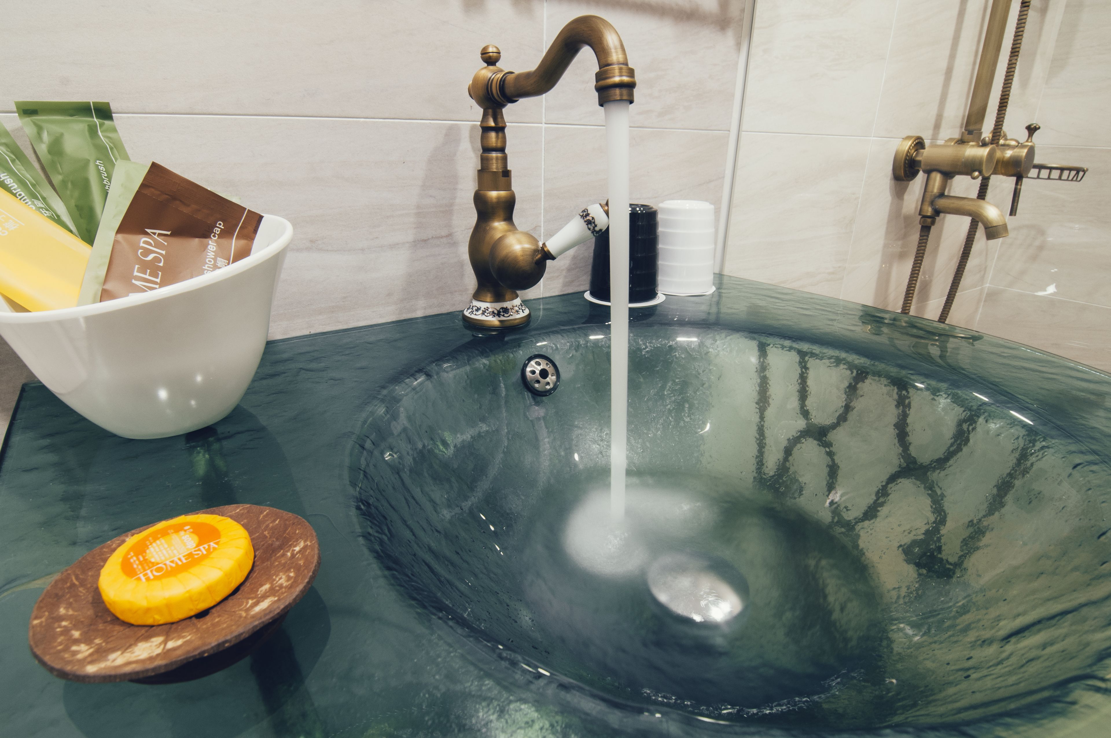 A bathroom sink with a glass basin, a white bowl with spa products, and a shower head.