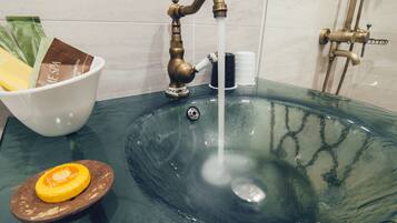 A bathroom sink with a glass basin, a white bowl with spa products, and a shower head.
