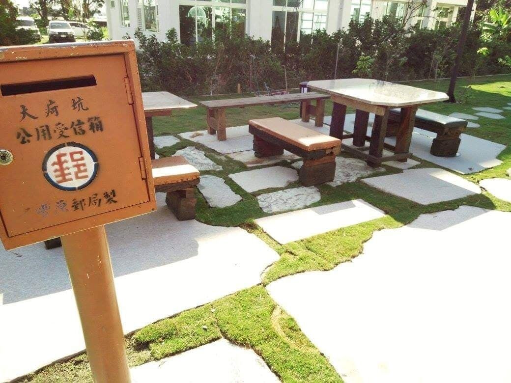 Une boîte aux lettres publique ornée de caractères chinois, une table de pique-nique et des bancs, le tout installé dans un espace aménagé avec des dalles de pierre et des zones d’herbe.