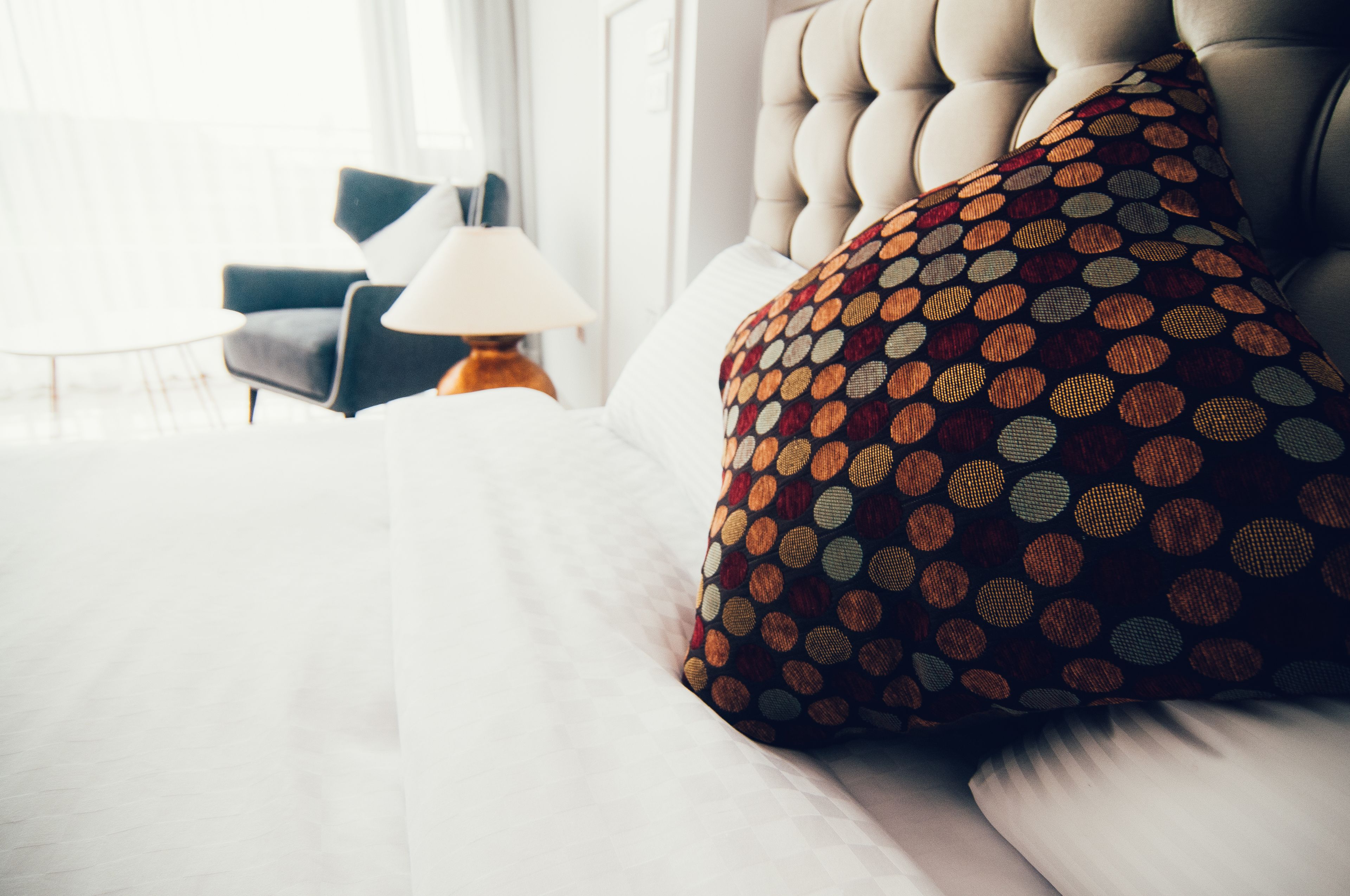 A neatly made bed with a patterned pillow, a lamp, and a chair in a well-lit room.