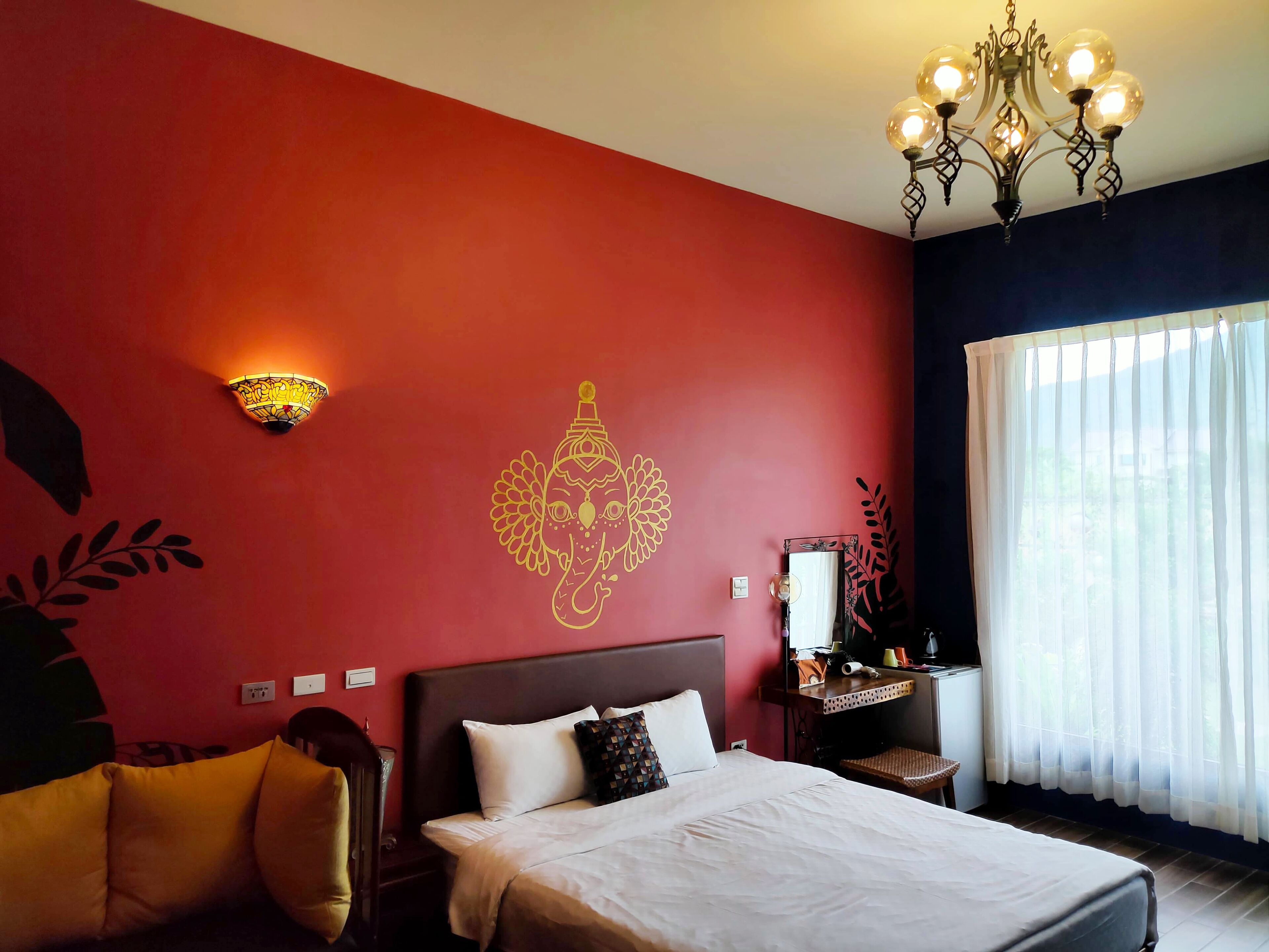 Une chambre d’hôtel avec un mur rouge, un lit avec une literie blanche, un lustre et une fenêtre ornée de rideaux légers.