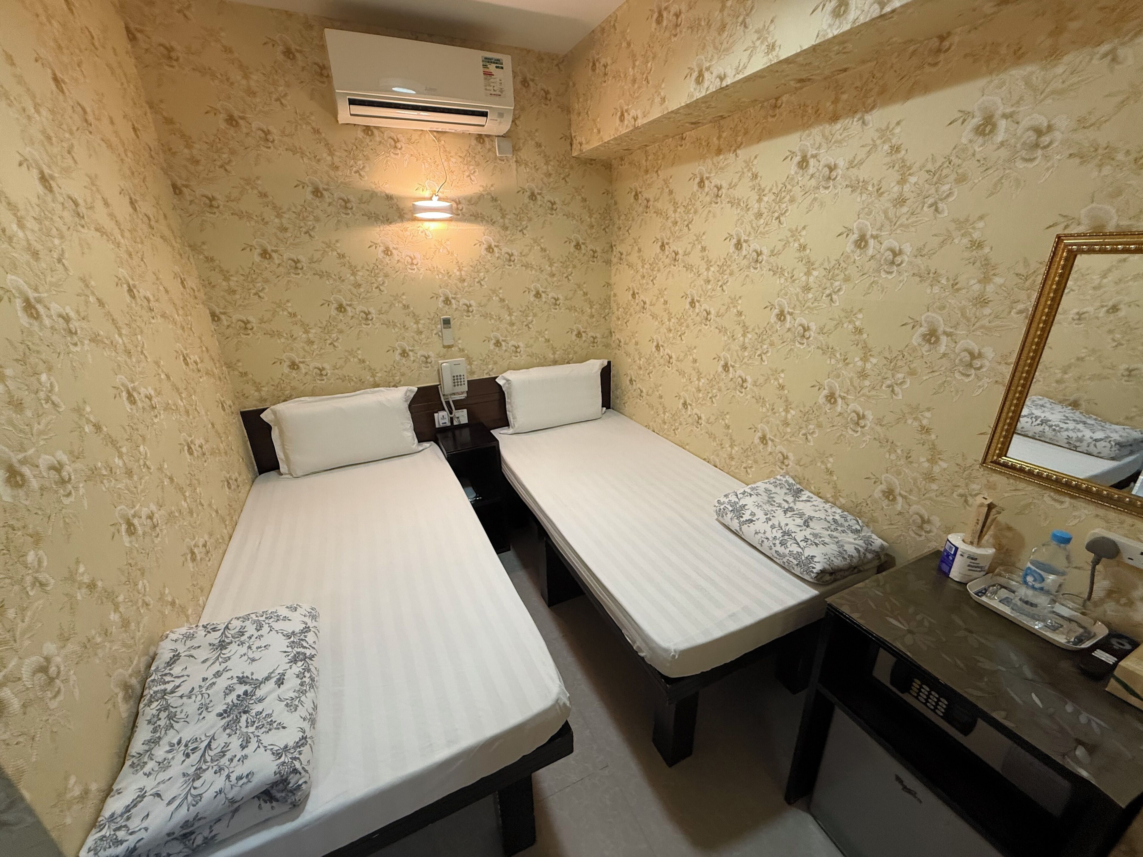 A room with two single beds, a small table with a mirror, and an air conditioning unit on the wall.