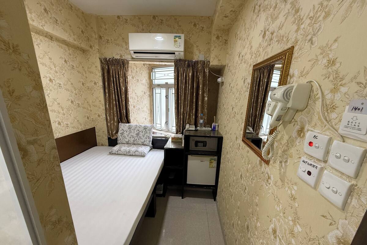 Une chambre d’hôtel compacte comprenant un lit, un bureau avec un réfrigérateur, un climatiseur, un sèche-cheveux et un miroir.