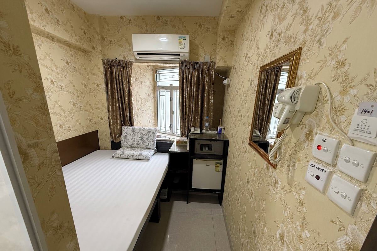 Habitación de hotel compacta con cama, escritorio con refrigerador, aire acondicionado, secador de pelo y espejo.