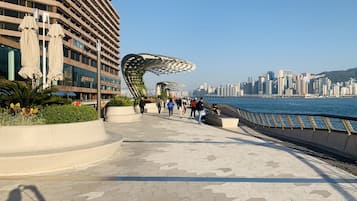 A waterfront promenade with a modern building, a curved metal structure, and a city skyline in the background.