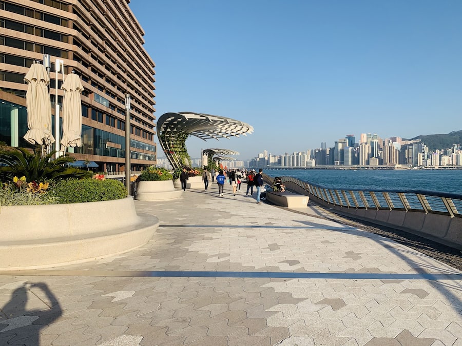 A waterfront promenade with a modern building, a curved metal structure, and a city skyline in the background.