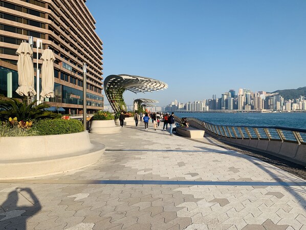 A waterfront promenade with a modern building, a curved metal structure, and a city skyline in the background. - Modern (Kowloon)