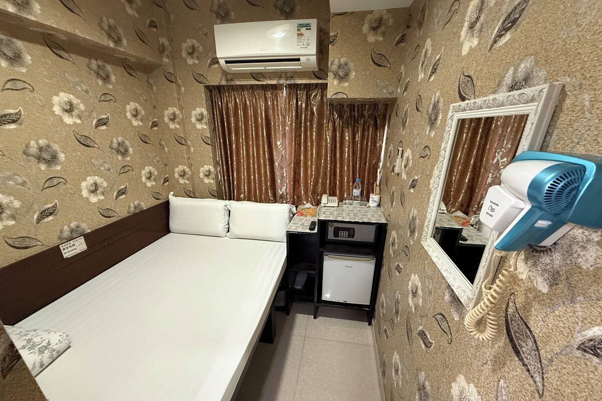Une chambre d’hôtel de taille réduite, comprenant un lit, un petit bureau avec un téléphone, un climatiseur, un mini-réfrigérateur et un miroir avec un sèche-cheveux.