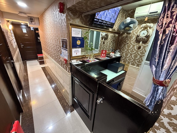 A hotel reception area with a reception desk, a television screen displaying a menu, a clock, and a mirror.